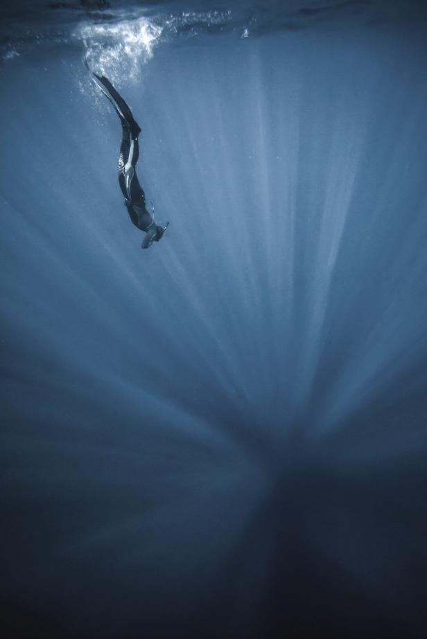 Ein Freitaucher taucht in tiefblaues Wasser mit Sonnenstrahlen ein.