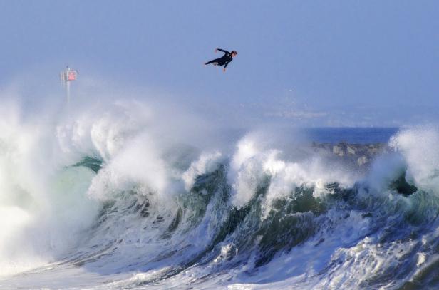 Ein Surfer springt über eine riesige Welle vor einem blauen Himmel.