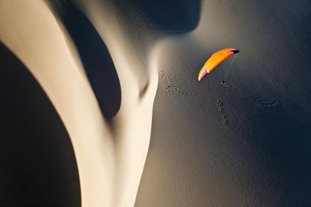 Ein orangefarbener Gleitschirm fliegt über eine Sanddüne.