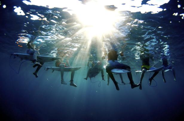 Eine Gruppe von Surfern sitzt auf ihren Brettern unter der Wasseroberfläche, während Sonnenstrahlen durch das Wasser scheinen.
