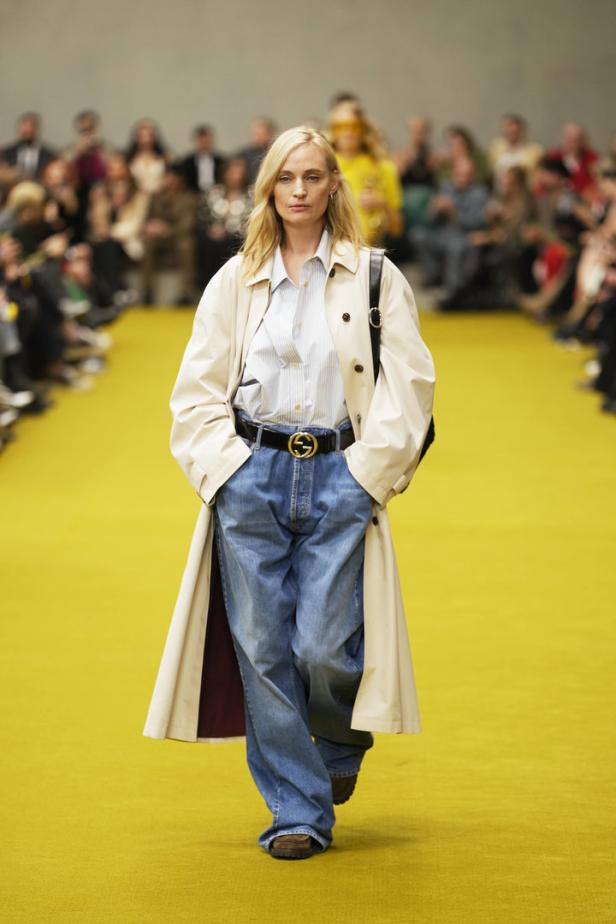 Eine Frau in einem Trenchcoat und Baggy Jeans läuft über einen gelben Laufsteg.