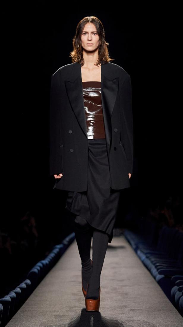 Eine Frau in einem schwarzen Blazer, einem braunen Lackleder-Top und einem schwarzen Rock auf einem Laufsteg.