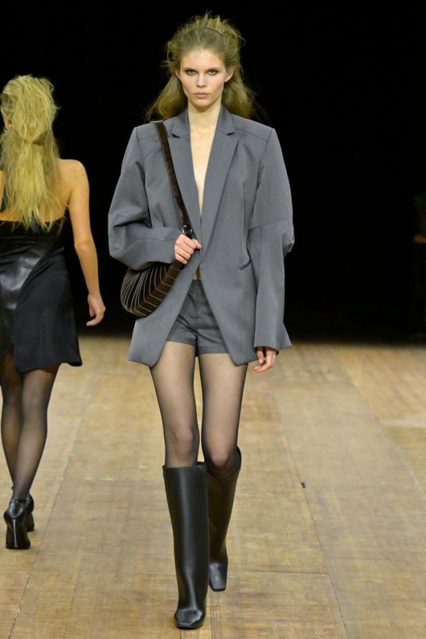 Eine Frau in einem grauen Blazer, Shorts, Strumpfhosen und Stiefeln auf einem Laufsteg.
