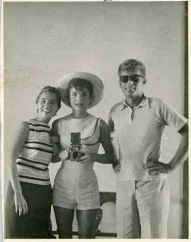 Jackie Kennedy mit einer Kamera, flankiert von einer Frau und einem Mann mit Sonnenbrille.