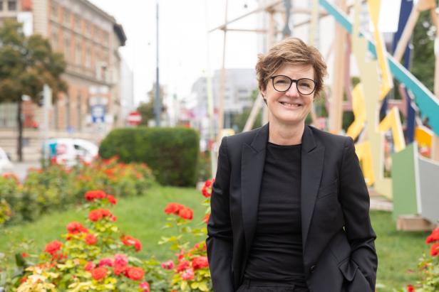 Eine Frau mit Brille und schwarzem Blazer steht vor einem Blumenbeet in der Stadt.
