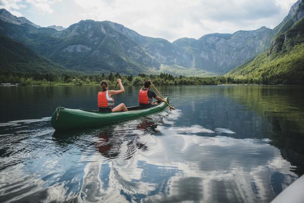 Zwei Personen paddeln in einem grünen Kanu auf einem See mit Bergen im Hintergrund.