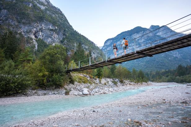 Zwei Wanderer überqueren eine Hängebrücke über einen türkisfarbenen Fluss in einer Berglandschaft.