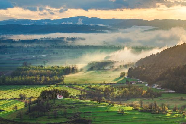 Eine grüne Landschaft mit Feldern, Wäldern und einer kleinen Kirche im Morgenlicht.