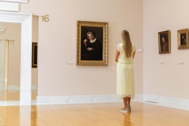 Eine Frau betrachtet ein Porträt einer älteren Dame mit einer Tasse in einem Museum.