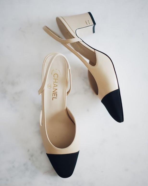 Zwei elegante Slingpumps von Chanel in Beige und Schwarz liegen auf einem Marmortisch.