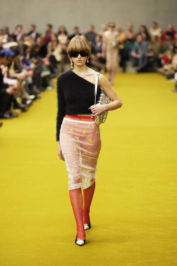Ein Model läuft auf dem Laufsteg in einem schwarzen Oberteil und einem transparenten Rock mit roten Strumpfhosen.