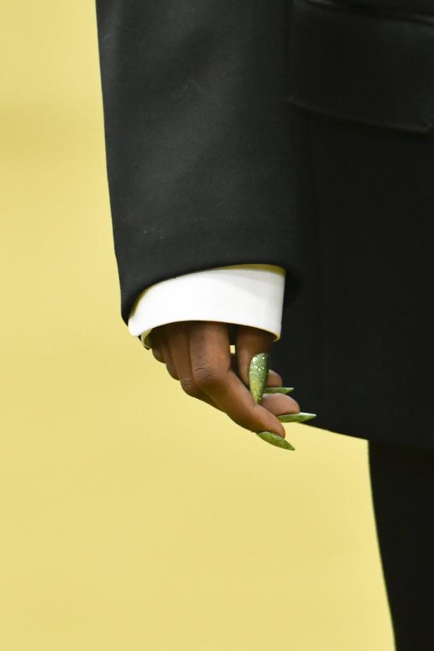 Die Hand einer Person mit langen, glitzernden, grünen Nägeln vor einem gelben Hintergrund.