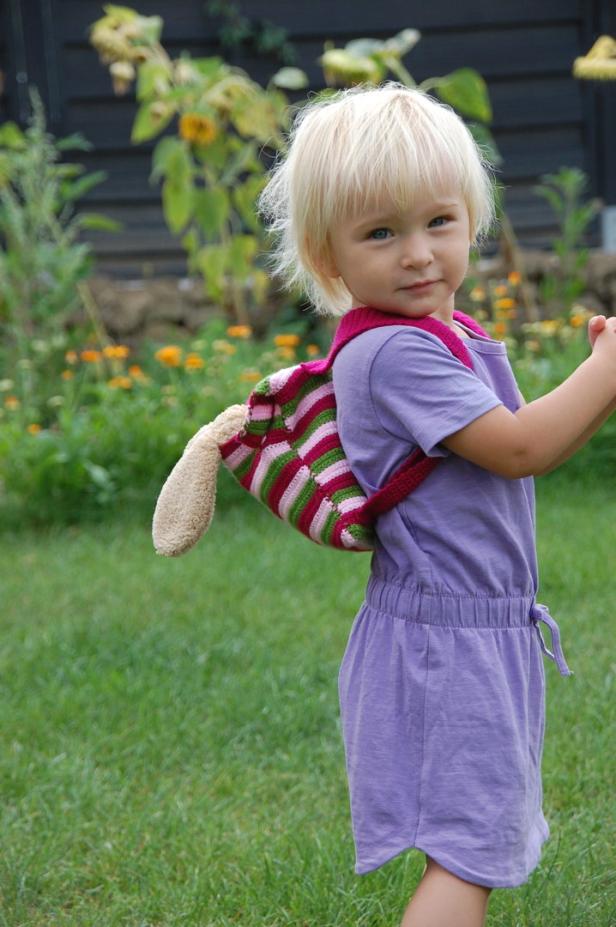Ein kleines Mädchen mit blonden Haaren und einem Hasenrucksack steht im Garten.