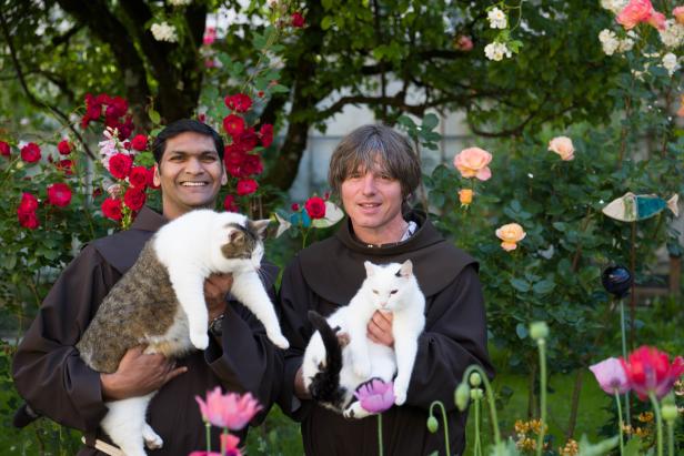 Zwei Männer in Kutten halten jeweils eine Katze in einem blühenden Garten.