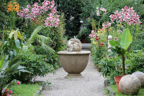 Ein Gartenweg führt zu einem Brunnen mit tropisch bemalter Kugel zwischen Blumenbeeten.