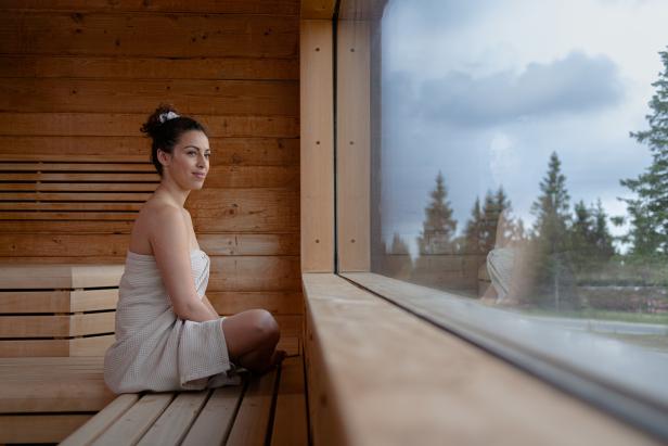 Eine Frau sitzt in einem Handtuch gehüllt in einer Sauna und blickt aus dem Fenster.
