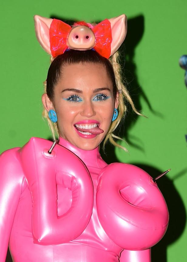 Miley Cyrus in einem pinkfarbenen Schweinekostüm mit passendem Stirnband und blauen Akzenten.