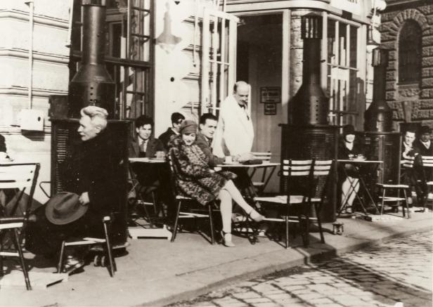 Menschen sitzen an Tischen vor einem Café mit Heizstrahlern.