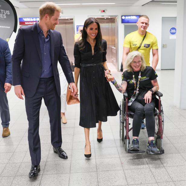 Prinz Harry und Meghan halten Händchen mit einer Rollstuhlfahrerin.