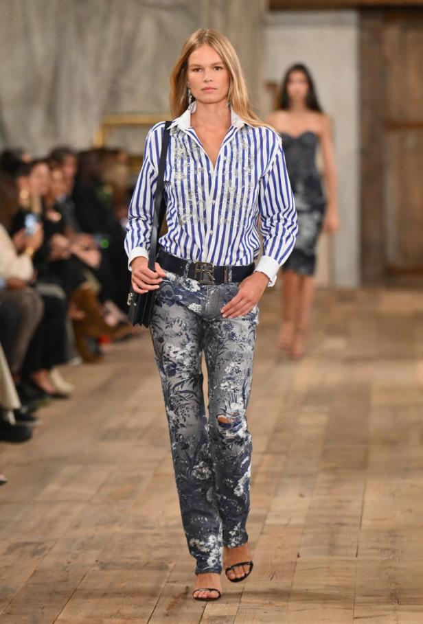 Ein Model läuft auf dem Laufsteg mit einer blau-weiß gestreiften Bluse und Jeans mit Blumenmuster.