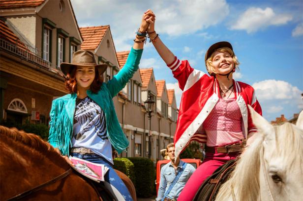 Zwei junge Frauen reiten auf Pferden und halten Händchen in einer Wohngegend.