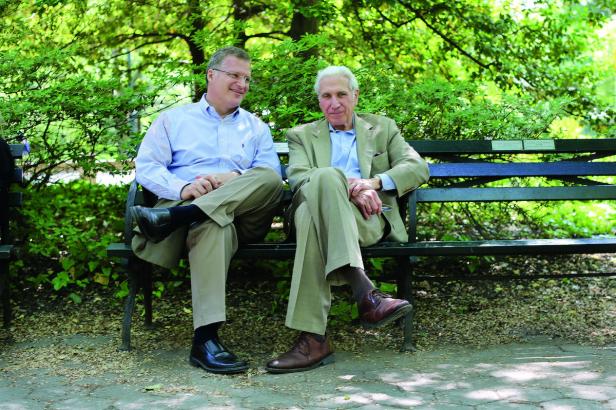 Zwei Männer sitzen auf einer Parkbank unter Bäumen.