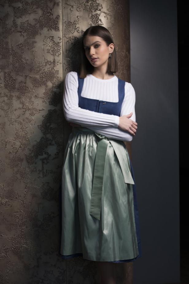 Eine junge Frau in einem Dirndl lehnt an einer verzierten Wand.