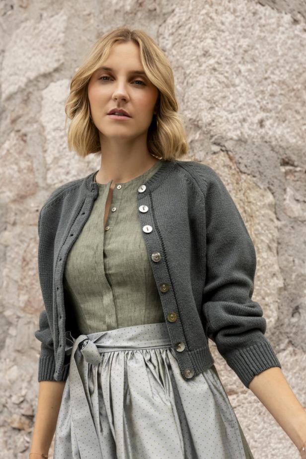 Eine Frau trägt eine Trachtenbluse, einen Rock und eine Strickjacke vor einer Steinwand.