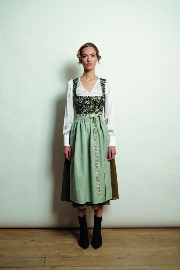 Eine Frau in einem traditionellen Dirndl mit grüner Schürze und braunen Stiefeln vor einem weißen Hintergrund.