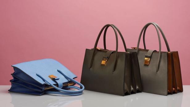 Drei Handtaschen in Blau, Grau und Olivgrün stehen vor einem rosa Hintergrund.