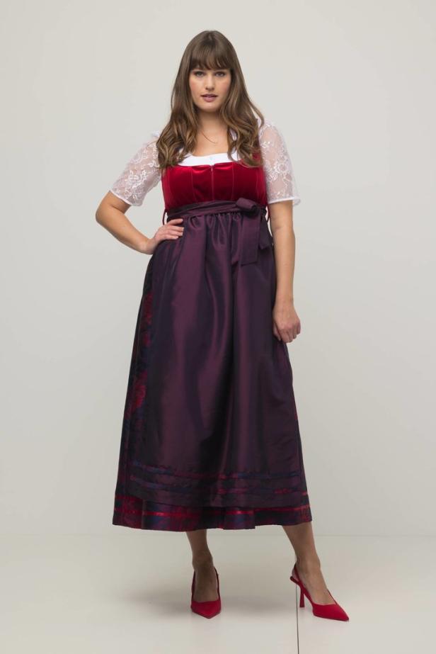 Eine Frau in einem roten Dirndl mit weißer Bluse und roten Schuhen posiert.