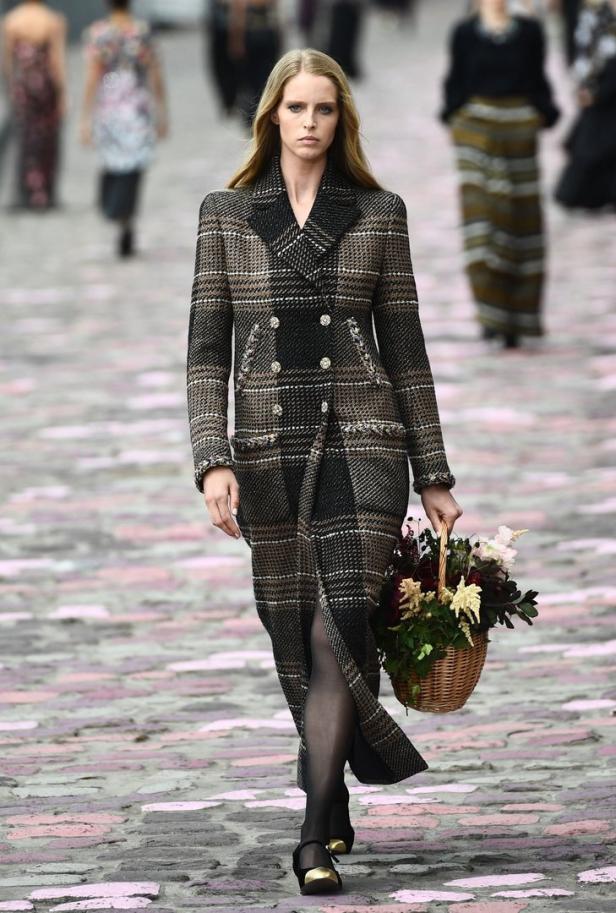Ein Model mit einem karierten Mantel und einem Blumenkorb auf einem Laufsteg.