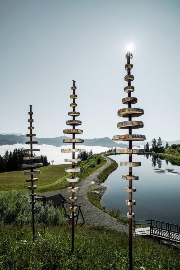 Drei Holzskulpturen stehen vor einem Bergsee in einer grünen Landschaft.