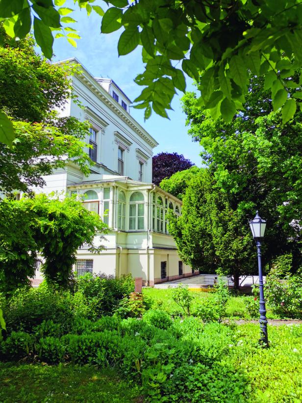 Ein weißes Gebäude mit einem Wintergarten, umgeben von üppigem Grün.