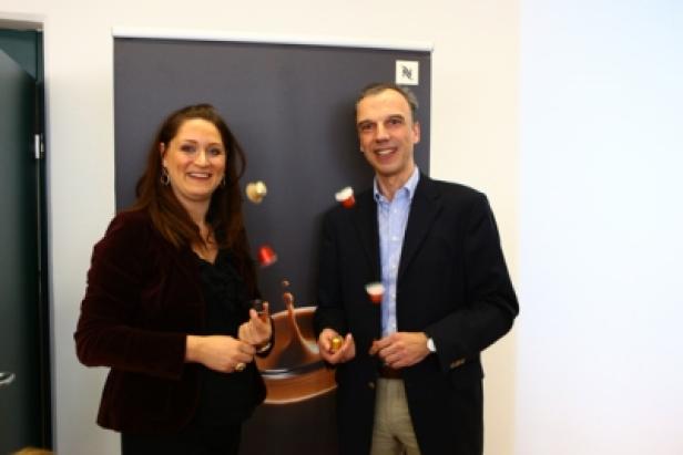 Eine Frau und ein Mann posieren vor einem Nespresso-Werbebanner und jonglieren mit Kaffeekapseln.