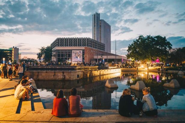 Menschen sitzen am Ufer eines Teiches vor der Stadthalle Chemnitz.