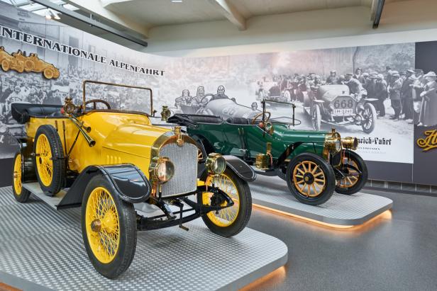 Zwei Oldtimer, ein gelber und ein grüner, stehen in einem Museum vor einer historischen Fotowand.