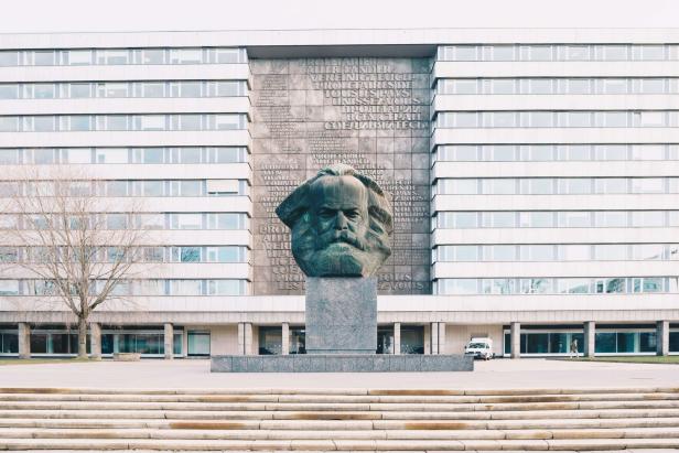 Die Karl-Marx-Büste in Chemnitz vor einem Gebäude mit dem Aufruf „Proletarier aller Länder, vereinigt euch!“.