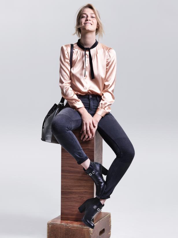 Eine Frau sitzt auf einem Holzblock und trägt eine rosa Bluse, Jeans und Stiefel.