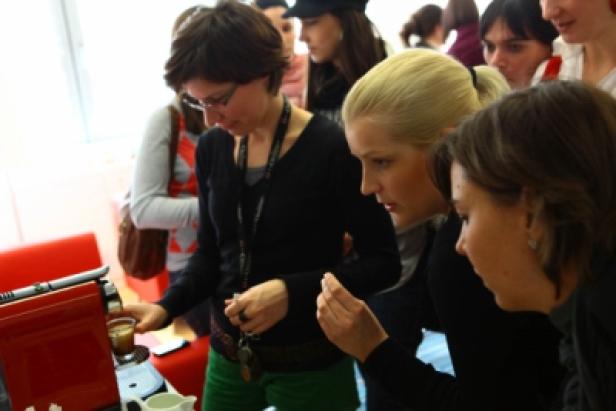 Eine Gruppe von Frauen beobachtet eine Frau, die eine Tasse Kaffee aus einer Kaffeemaschine zubereitet.