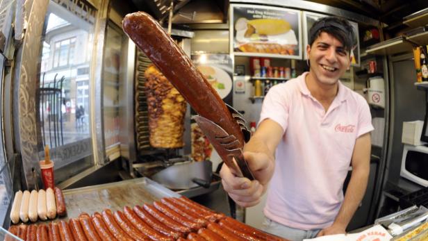 Ein Imbissverkäufer hält eine riesige Wurst mit einer Zange in die Kamera.