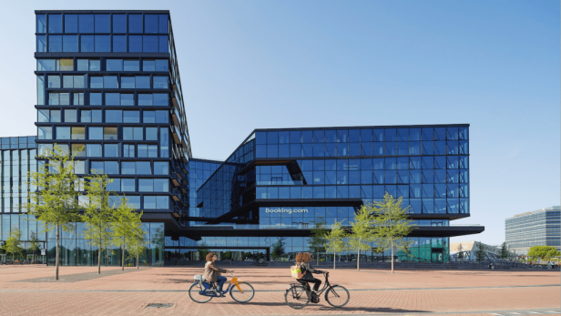 Zwei Personen fahren vor dem modernen Bürogebäude von Booking.com mit Fahrrädern.