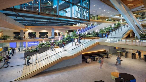 Mehrere Personen nutzen Treppen und Rolltreppen in einem modernen, begrünten Bürogebäude.