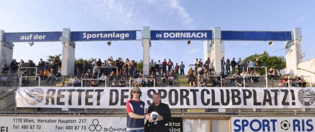 Zwei Männer stehen vor einer Tribüne mit dem Banner „Rettet den Sportclubplatz!“.