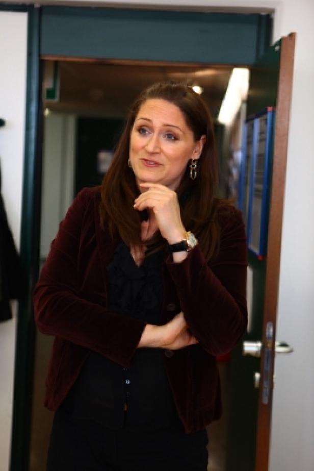 Eine Frau in einem roten Blazer steht nachdenklich vor einer Tür.