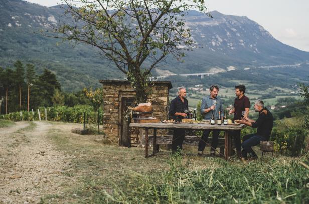 Vier Männer stehen im Freien an einem Tisch mit Wein und Speisen vor einer Berglandschaft.