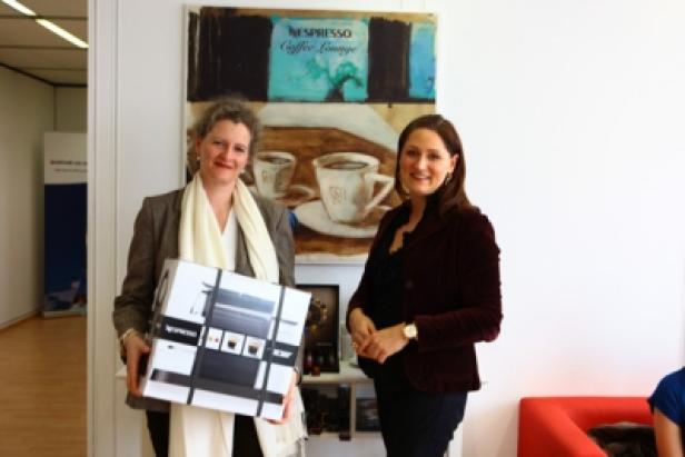 Zwei Frauen posieren mit einer Nespresso-Maschine vor einem Bild mit Kaffeetassen.