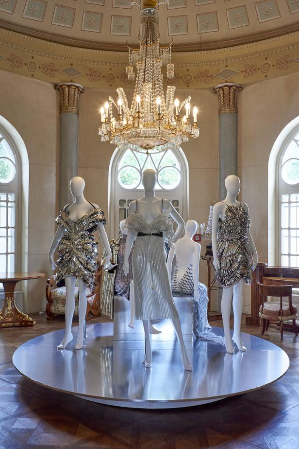 In einem Ausstellungsraum stehen mehrere Mannequins mit aufwändigen, silberfarbenen Kleidern unter einem Kronleuchter.