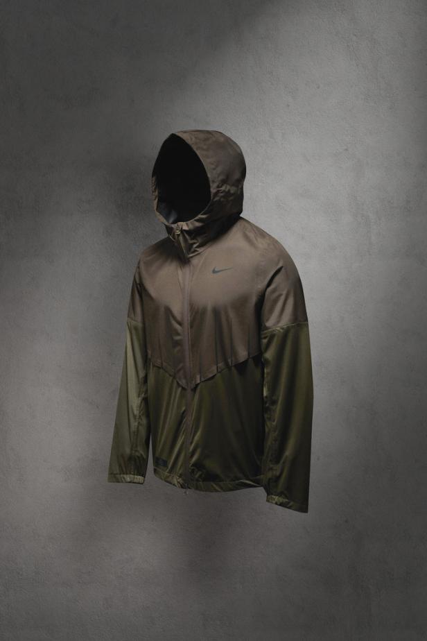 Eine olivgrüne Nike Windrunner Jacke mit Kapuze vor einem grauen Hintergrund.