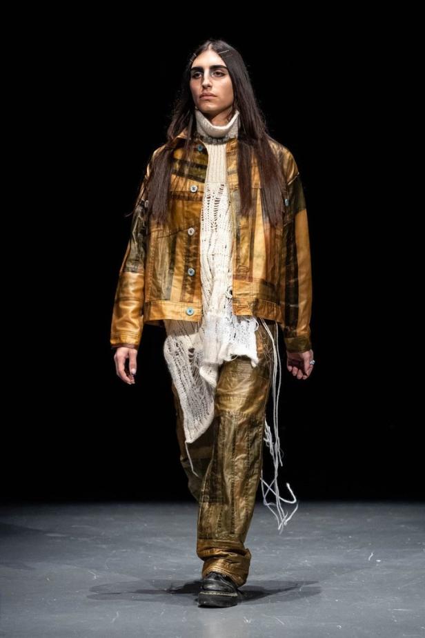 Ein Model präsentiert ein Outfit mit Jacke, Pullover und Hose in Goldtönen auf einem Laufsteg.